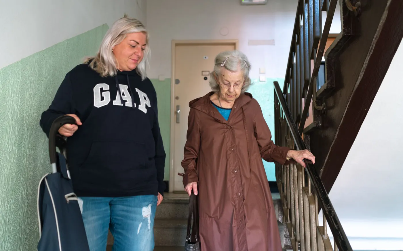 Une dame âgée descend l’escalier de son immeuble, accompagnée