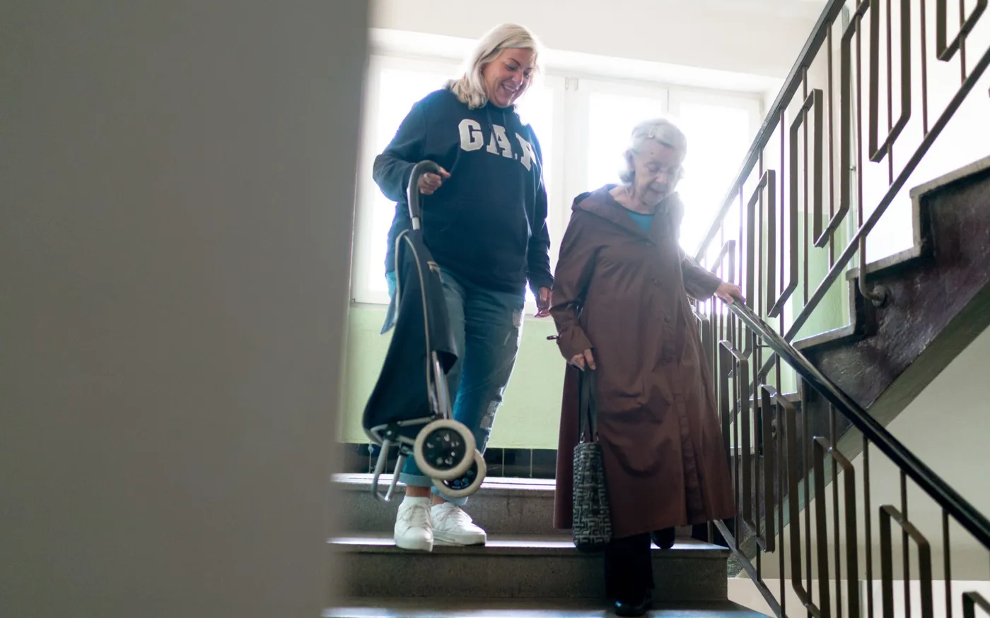 Une aide à domicile accompagne une dame âgée qui descend l’escalier de son immeuble